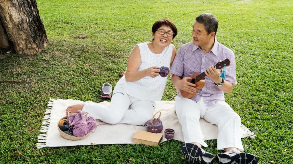 A joyful older Asian couple sits on a blanket in a park, with the man playing a ukulele and the woman holding a mug and laughing.