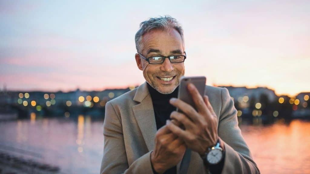 A smiling, gray-haired man in a suit jacket looks at his phone outdoors at sunset.