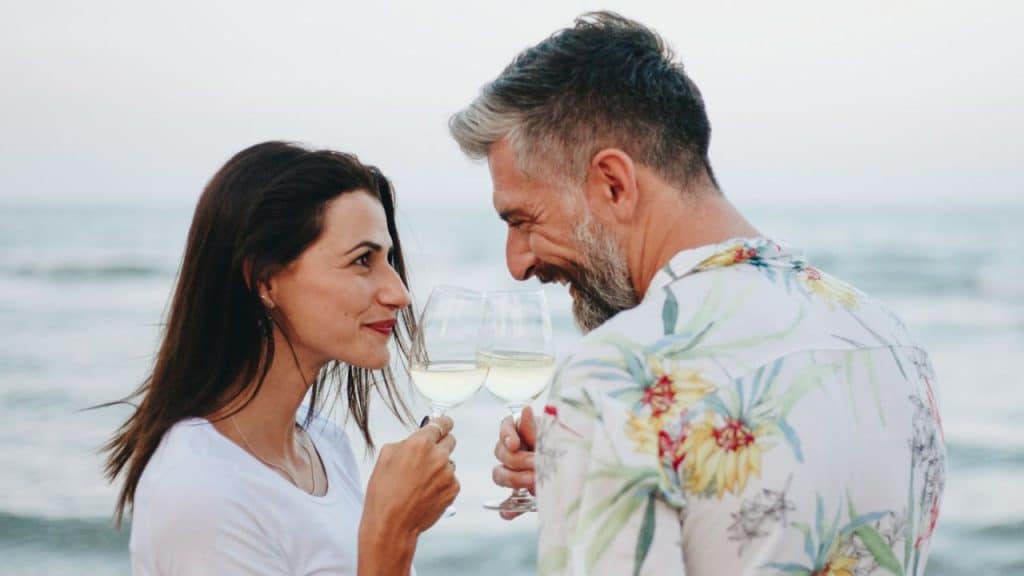 A smiling man and woman are clinking white wine glasses by the ocean.