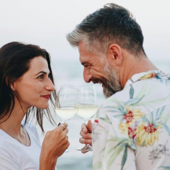 A smiling man and woman are clinking white wine glasses by the ocean.
