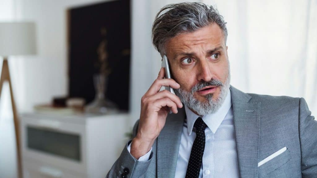 A confused or concerned middle-aged man in a suit is speaking on a mobile phone indoors.