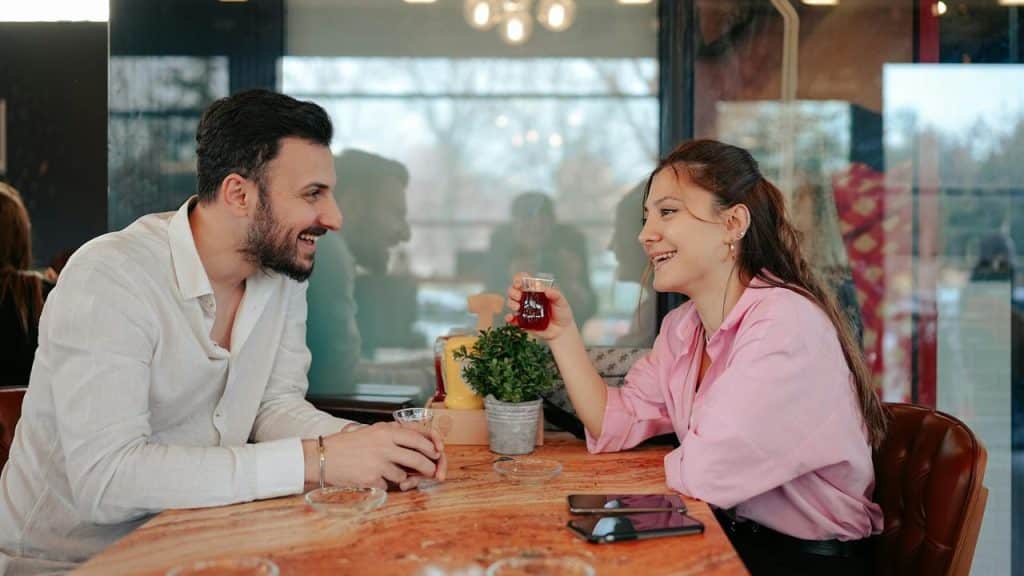 A man and woman smiling at each other while holding drinks at a café table.