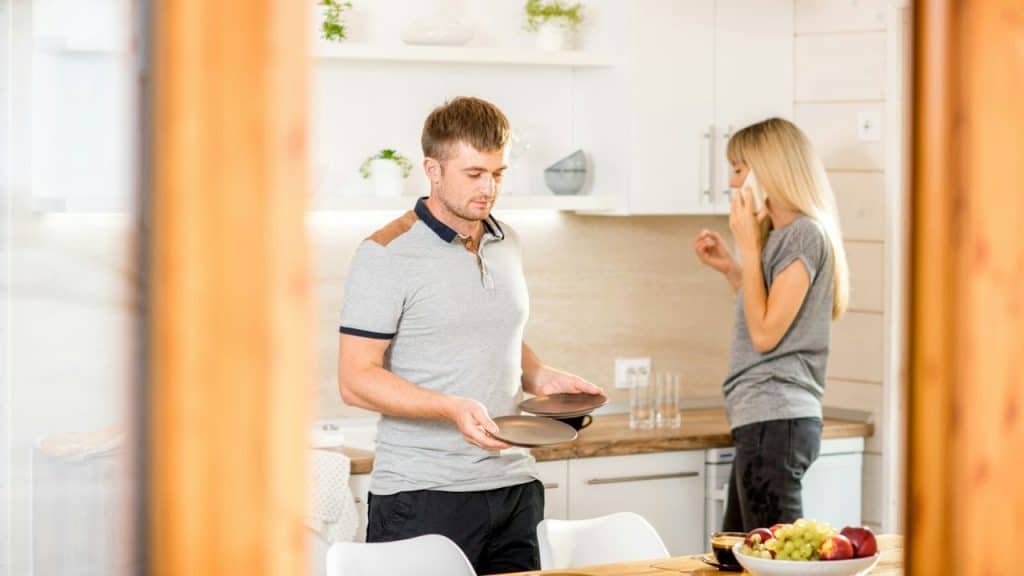 A man holds plates at a table while a woman talks on her phone in the kitchen behind him.