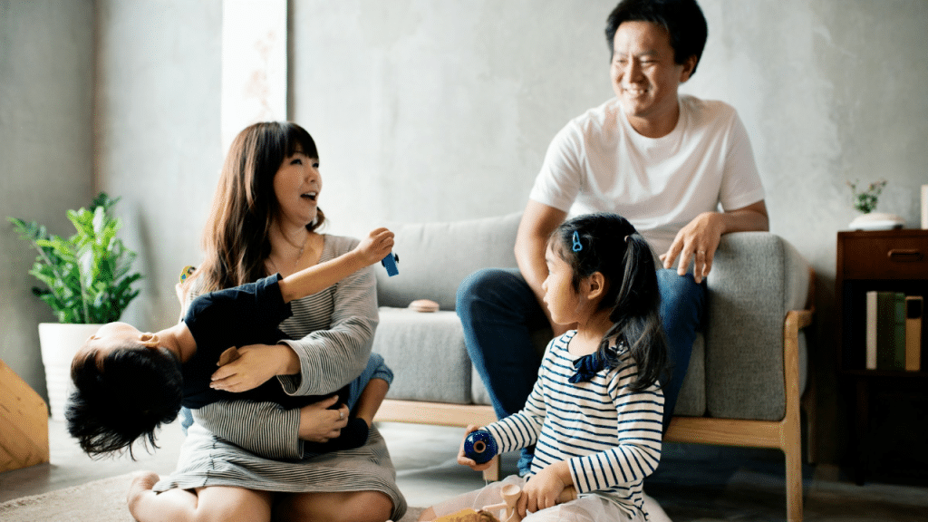 A man smiles while sitting on a couch, watching his wife play with their two children.