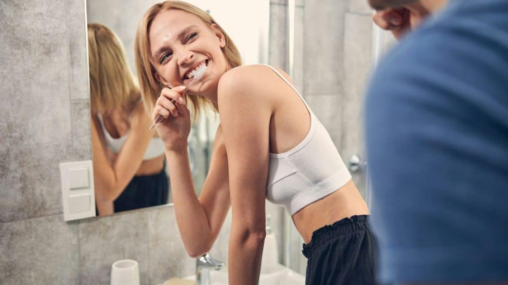 A happy blonde woman in a white tank top is **brushing her teeth** in a bathroom, looking directly at the camera.