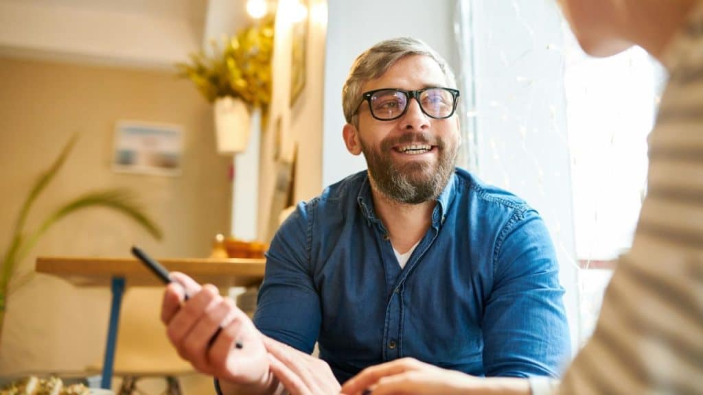 A smiling, bearded man wearing glasses and a blue shirt talks indoors.