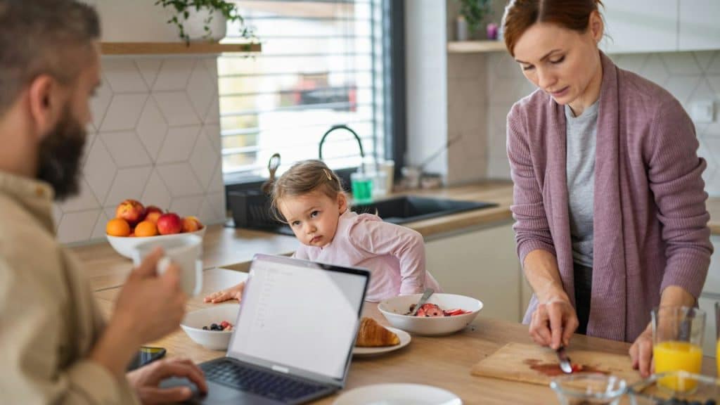 A family is in a kitchen; a man works on a laptop, a woman cuts food, and a girl watches.