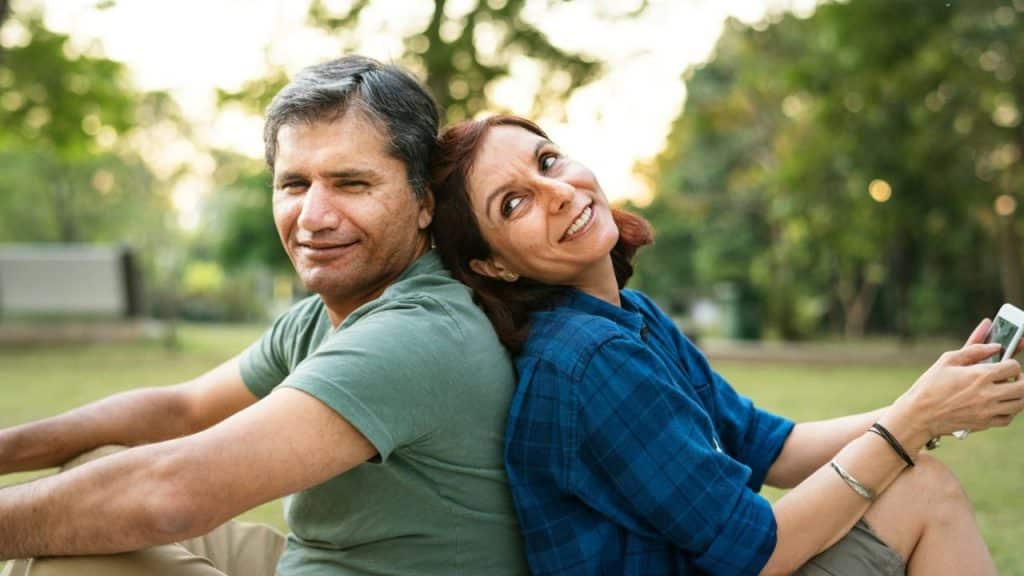 A middle-aged man and woman are sitting back-to-back in a park, smiling at the camera.