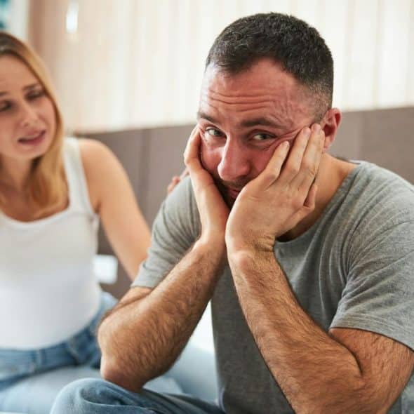 A worried woman touches the back of a stressed man's shoulder.