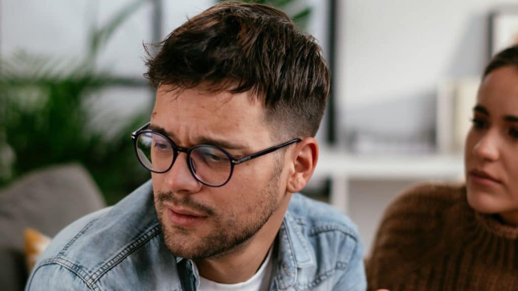 A man wearing glasses looks away from a woman, who is partially visible.