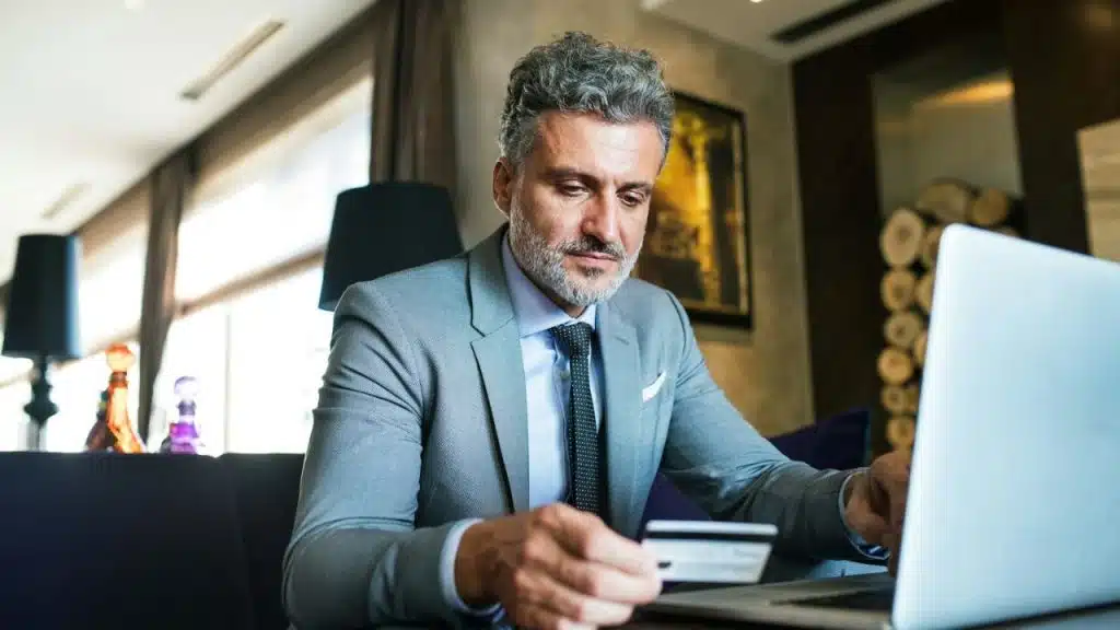A man in a gray suit sits in a chair, holding a credit card and looking down at a laptop.
