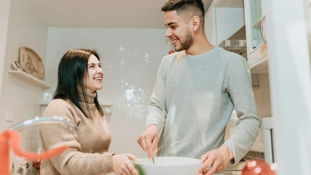 A smiling man and woman are cooking together in a bright kitchen.