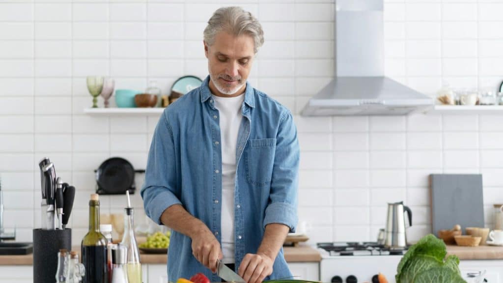 A handsome, mature man in a denim shirt is chopping vegetables in a clean, modern kitchen.