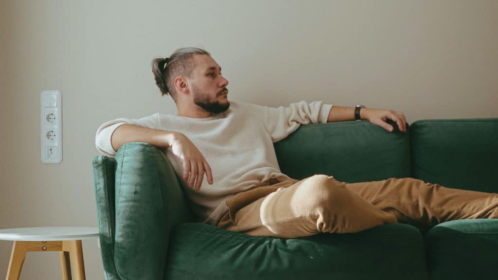 A man with a beard and hair pulled back sits on a green couch, looking to the right.