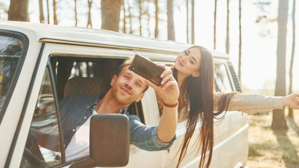 A smiling couple in a white van takes a selfie in a sunlit forest.