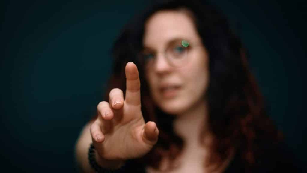 A woman wearing glasses with her face blurred out holds her index finger up toward the camera.