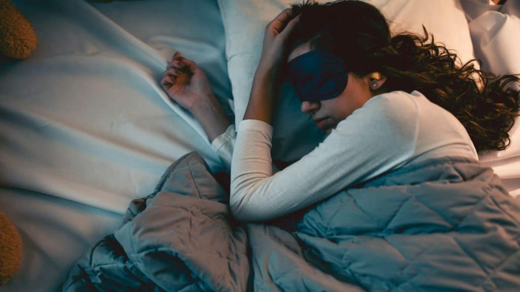 A person with long, dark hair is sleeping on their side in bed with a sleep mask on.