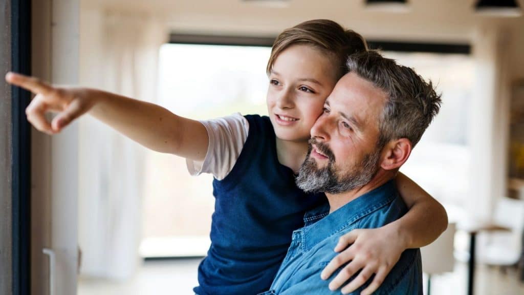 A man and a child look out a window; the child points while hugging the man.