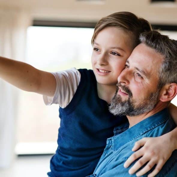 A man and a child look out a window; the child points while hugging the man.