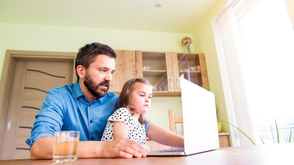 A father and his young daughter sit at a table looking at a laptop computer.