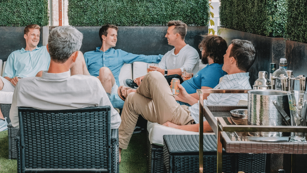A group of men sit and talk on an outdoor patio, with one man holding a can of beer.