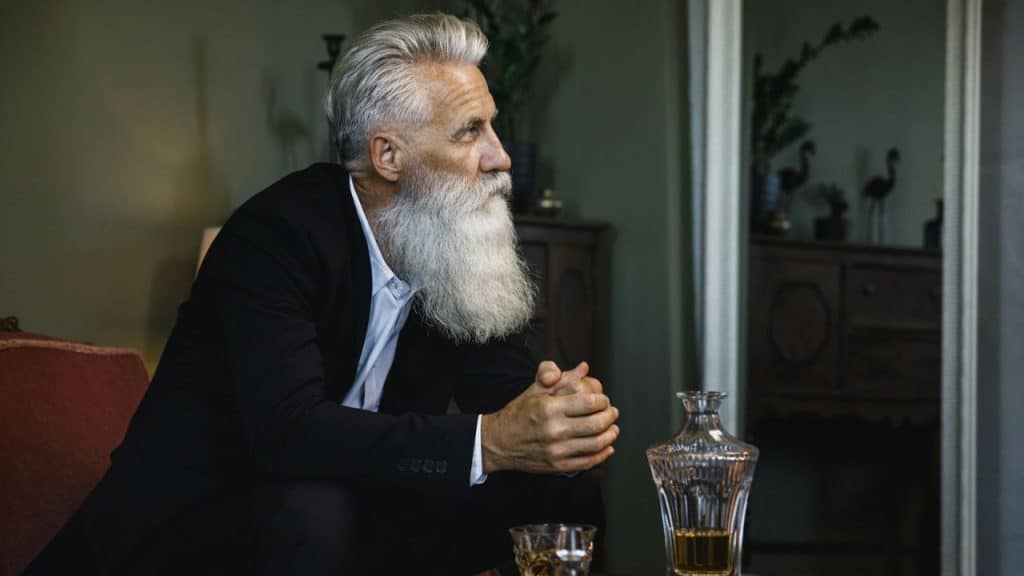 A man with a large white beard and black suit sits in an armchair, looking to the side.
