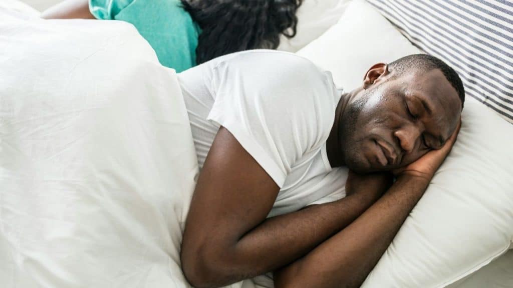 A man and a woman lie in bed. The man is sleeping on his side, facing the camera.