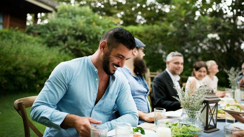 A man with a beard and a blue shirt laughs at a long table during an outdoor gathering.