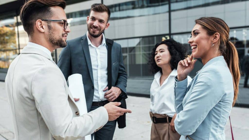 A group of four business professionals is standing outside, talking and smiling.
