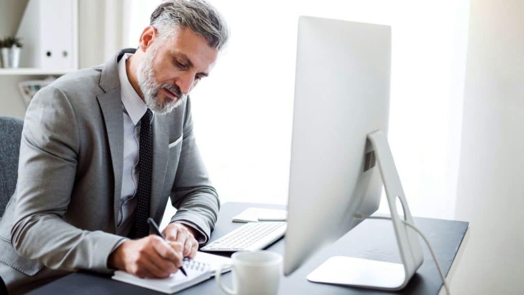 A middle-aged businessman with a beard is writing in a notebook at his desk.