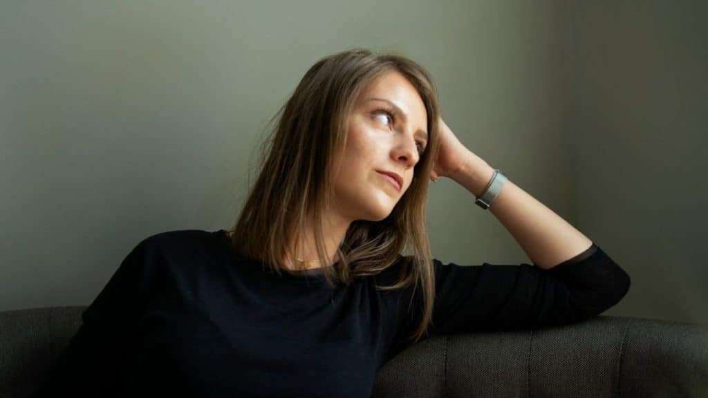 A woman with light brown hair sits on a couch, looking away and resting her head on her hand.