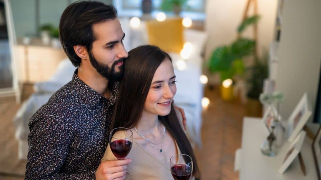 A bearded man and a woman stand together, holding two glasses of red wine indoors.