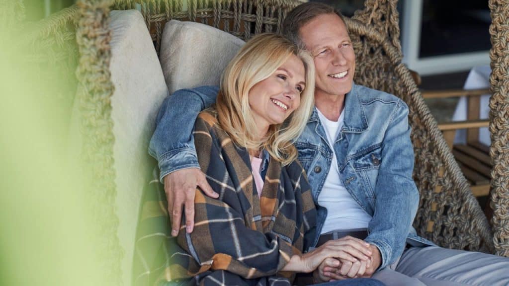A happy, older couple sits on a swinging chair, holding hands and smiling as they look into the distance.