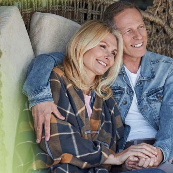 A happy, older couple sits on a swinging chair, holding hands and smiling as they look into the distance.