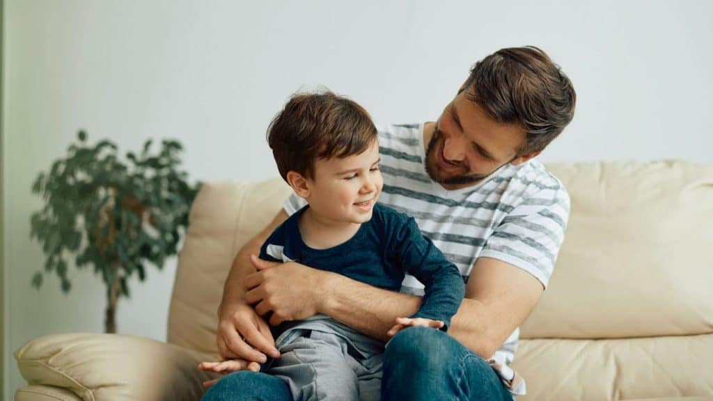 A father sits on a couch, hugging and smiling at his young son on his lap.