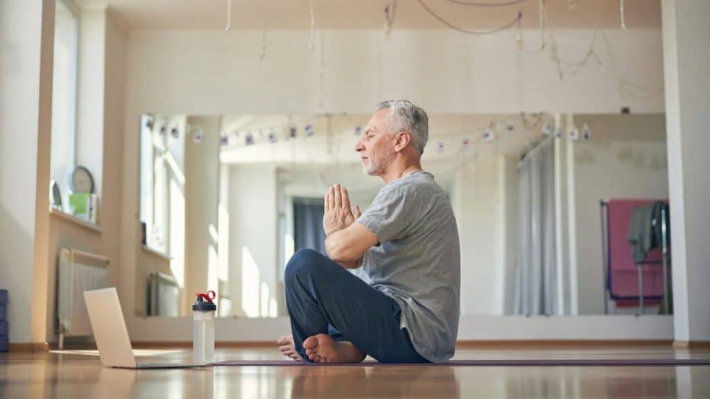 An older man with gray hair is sitting on a yoga mat in a meditation pose, with a laptop and water bottle in front of him.