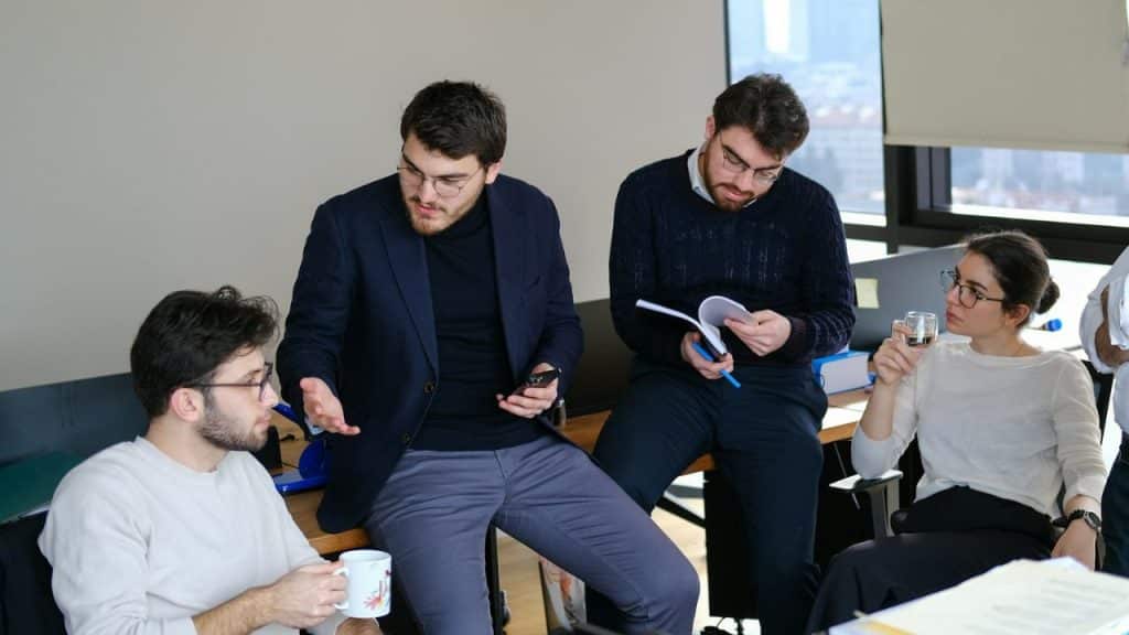 Four colleagues sit and stand around a desk in an office, engaged in a discussion.