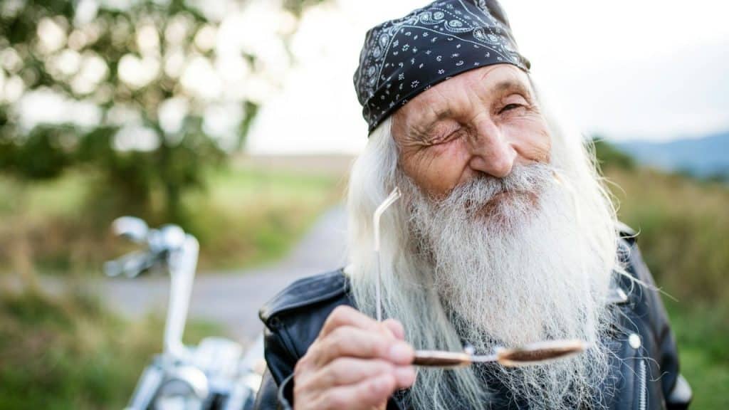 A smiling older man with a long white beard and a black bandana holds sunglasses.