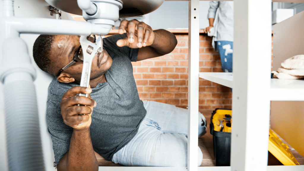 A man lies under a sink, using a wrench to fix the pipes, while a woman stands nearby.