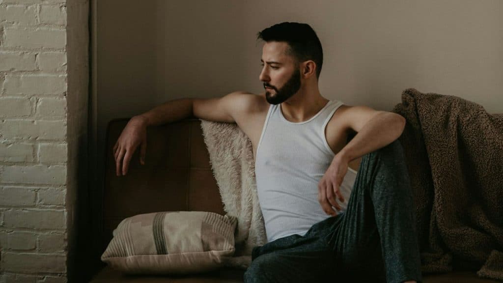 A man with a beard sits on a couch in a white tank top, looking to the side.