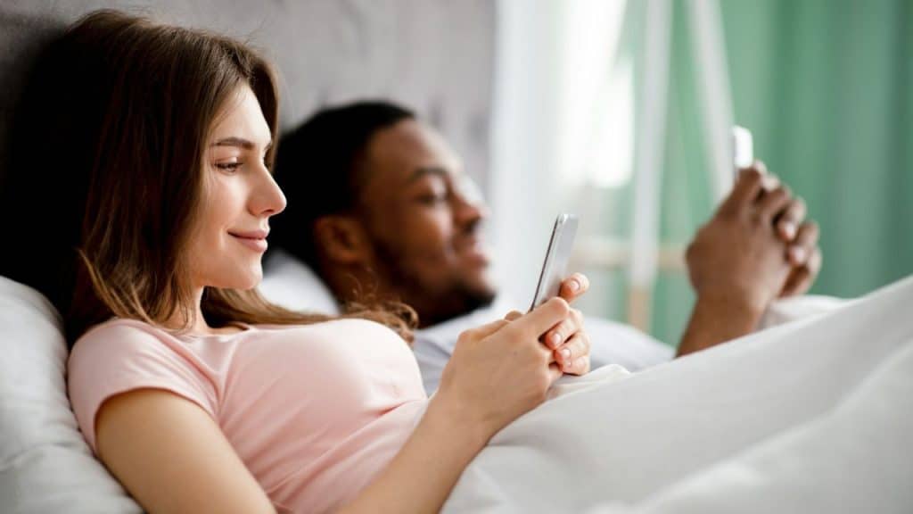 A white woman and Black man lie in bed, both looking at their phones.