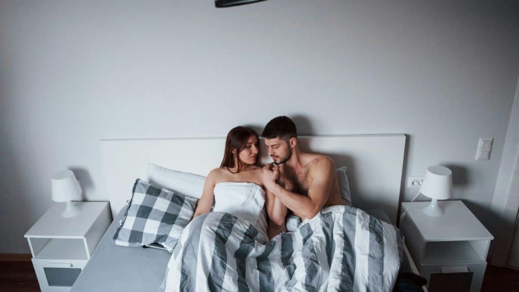 A smiling couple lies in a bed, holding hands and looking at each other.
