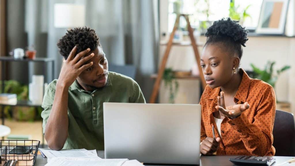 A stressed man holds his head in his hand while a woman gestures at a laptop.