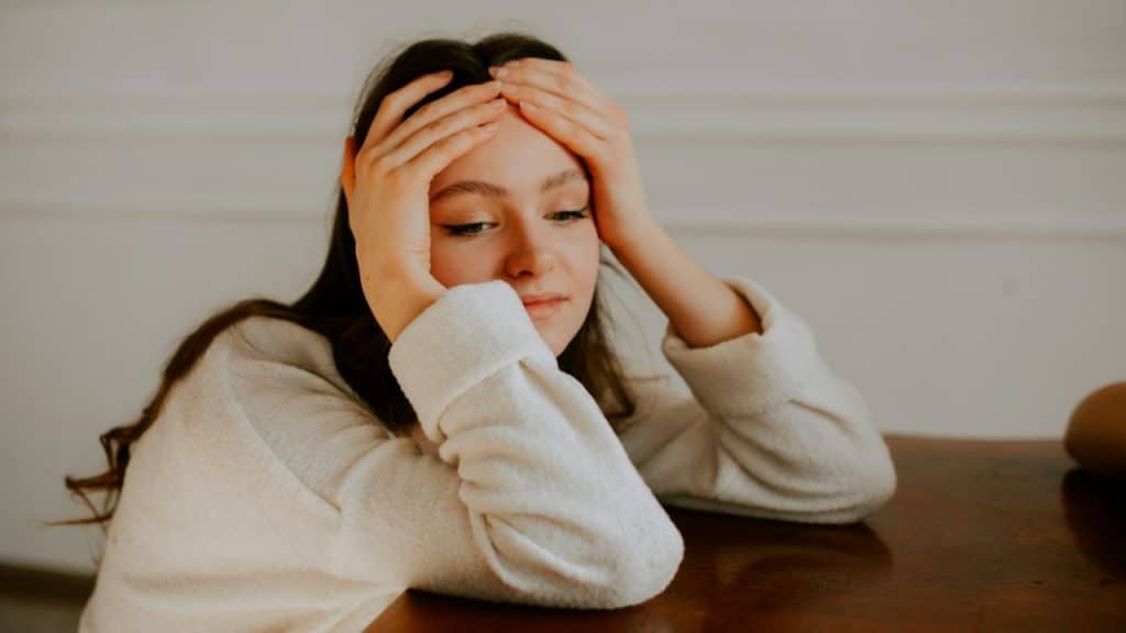 A young woman sits at a table with her head in her hands, looking down with a sad or pensive expression.
