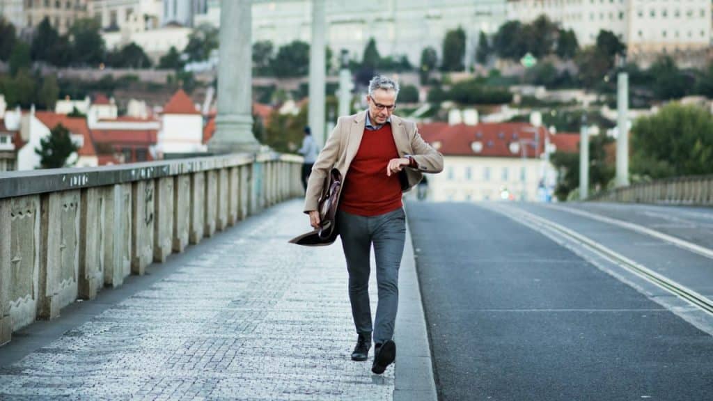 A busy businessman in a red sweater and blazer checks his watch while walking.