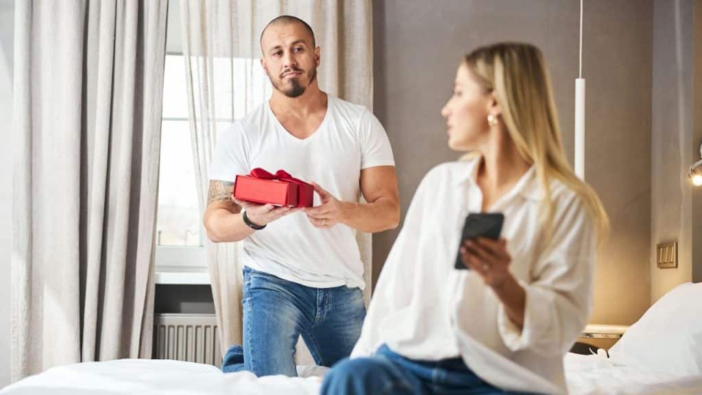 A man holding a wrapped gift kneels in front of a woman who is looking at her phone.