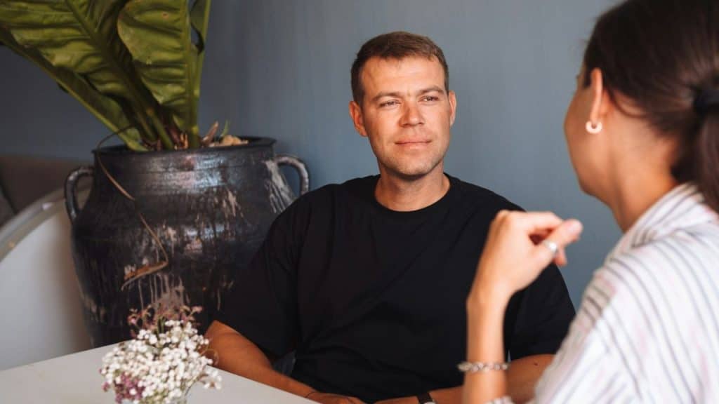 A man in a black shirt sits at a table, looking at a woman during a conversation.