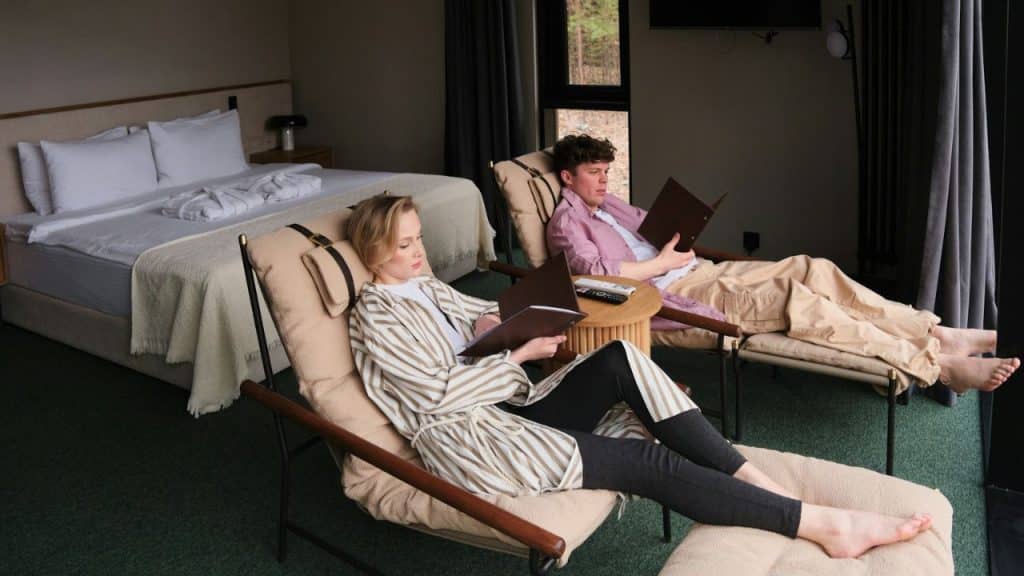 A man and a woman are relaxing in lounge chairs reading books in a hotel room.