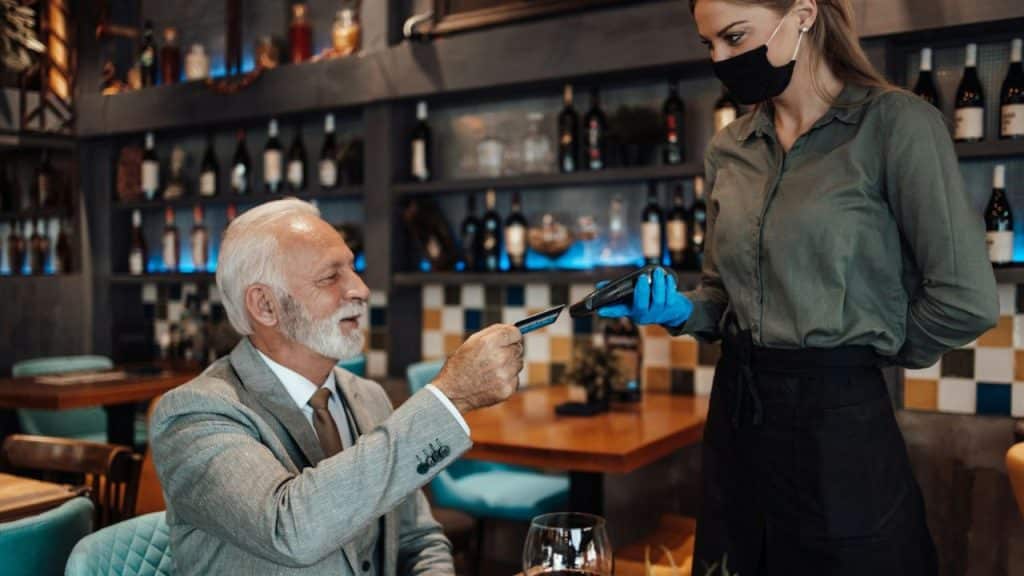 An older, well-dressed man is paying with a credit card to a waitress wearing a mask and gloves.