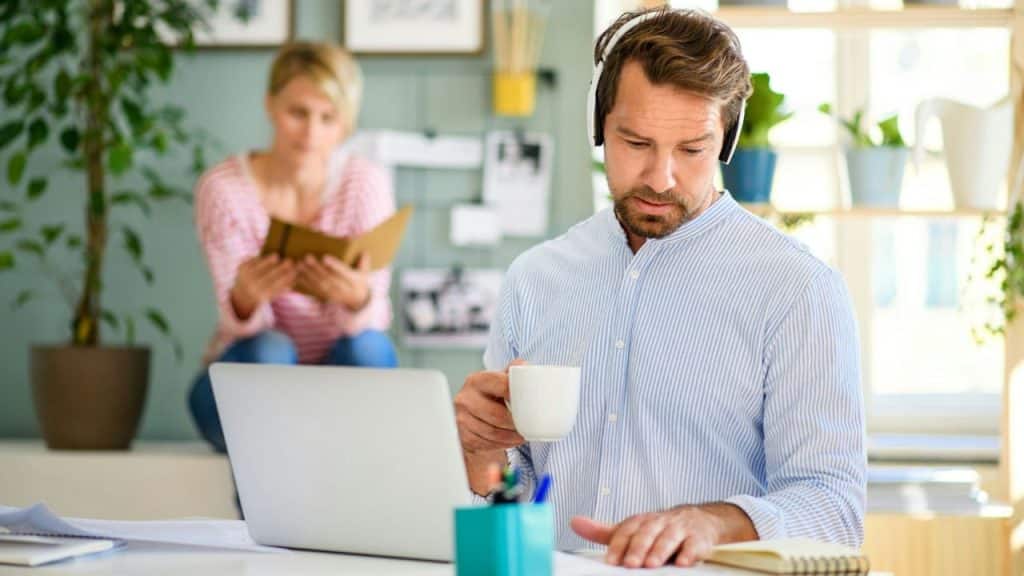 A man with headphones works on a laptop as a woman reads in the background.